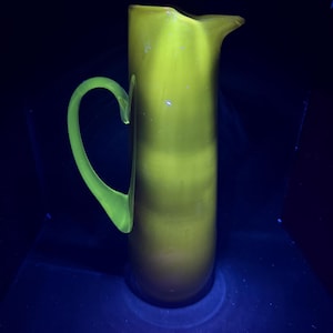 Puede incluir: Una jarra alta de vidrio amarillo verdoso con un asa curva y un pico. La jarra tiene un cuerpo liso y redondeado y un asa verde brillante contrastante. La jarra es probablemente vintage o antigua y es un artículo decorativo.
