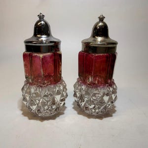 Puede incluir: Dos saleros y pimenteros antiguos. Cada uno tiene una base transparente facetada, una sección central de color rojo rubí y una tapa de metal plateado. Los saleros están sobre una superficie blanca.