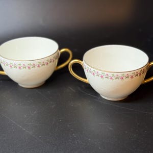 May include: Two white porcelain teacups with gold handles and rims. Each cup features a delicate floral pattern of pink flowers and green leaves around the rim. The cups are set against a dark background.