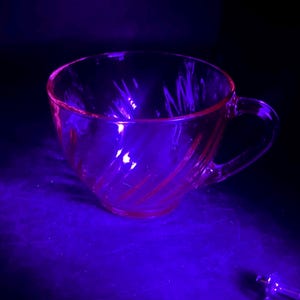 May include: A clear glass teacup with a handle. The cup has a spiral design and is illuminated with a purple light, creating a pink hue. The teacup is on a dark surface.