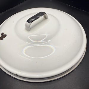 Può includere: Coperchio in smalto bianco con manico nero. Il coperchio ha una forma circolare con un centro leggermente rialzato e un bordo nero. C'è una piccola area di ruggine sulla superficie. Il coperchio è probabilmente per una pentola o una padella.