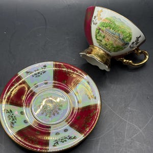 Könnte beinhalten: Ein Vintage-Tee-Set, bestehend aus einer Tasse und einer Untertasse. Die Tasse hat einen goldenen Henkel und Rand, mit einem Landschaftsbild und dem Text "Bühlertähe". Die Untertasse hat ein rot-goldenes Karomuster mit floralen Akzenten.