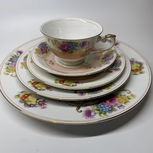 May include: A vintage tea set comprising a teacup, saucer, and two plates. The set displays a white base adorned with floral patterns in pink, blue, yellow, and purple. Gold trim edges the teacup and plates, adding a touch of elegance.