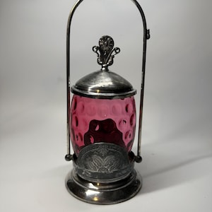 May include: A vintage, ruby-coloured glass jar with a silver-toned metal lid and base. The jar has a textured, dimpled surface and is held within a silver metal frame with a handle. The lid is topped with a decorative floral element.