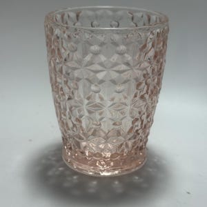 Puede incluir: Un vaso transparente teñido de rosa con un patrón geométrico texturizado. El vaso tiene una base corta y redondeada y una forma cilíndrica. El diseño presenta un motivo repetido de diamantes y puntos, creando una estética vintage.