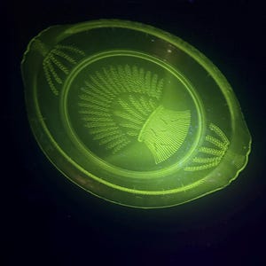 May include: A vintage, oval-shaped, green glass plate with a wheat sheaf design in the centre and along the edges. The plate has a scalloped rim and two small handles. The glass glows brightly against a dark background.