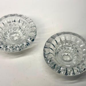 May include: Two clear, round glass candle holders. Each has a textured, ridged exterior and a central well for holding a candle. The glass is transparent. Suitable for home decor, these candle holders are a decorative item.