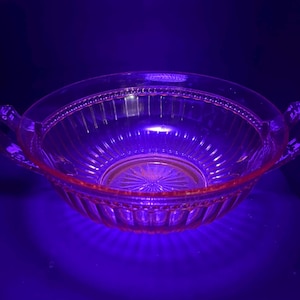 Könnte beinhalten: Eine transparente, rosa Glasschale mit zwei Griffen. Die Schale hat ein geripptes Design an den Seiten und ein dekoratives Muster am Boden. Der Rand ist mit kleinen, erhabenen Punkten versehen. Die Schale ist wahrscheinlich Vintage oder antik.