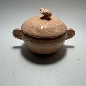 May include: A pale pink ceramic sugar bowl with a matching lid featuring a sculpted finial. The bowl has two side handles and a raised, swirling relief pattern covering both the lid and the body, finished with a glossy, smooth glaze throughout.