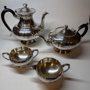 May include: A silver tea set comprising two teapots, a creamer, and a sugar bowl. The teapots feature black handles and lids. The creamer and sugar bowl have matching handles. The set is presented on a white surface.