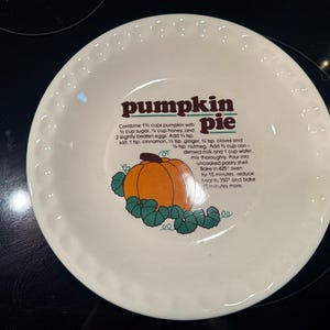 May include: White ceramic pie plate with a decorative pumpkin pie design. The plate features a pumpkin illustration with green leaves and the words "pumpkin pie" in brown text. Recipe instructions are also printed on the plate.