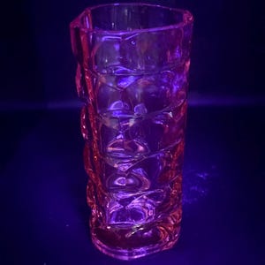 May include: A tall, clear glass vase with a textured, geometric design. The vase has a slightly flared top and is illuminated with pink and purple lighting. The vase is likely used for decorative purposes. The design is reminiscent of Art Deco.