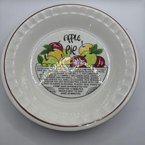 Puede incluir: Plato de cerámica blanco para pastel con borde marrón y diseño decorativo de tarta de manzana. El plato presenta las palabras "APPLE PIE" y un gráfico de manzanas. Incluye instrucciones para hornear tarta de manzana.