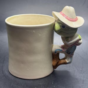 May include: Cream ceramic mug with a novelty frog handle. The frog is wearing a cowboy hat with a pink band and a blue shirt. The mug is cylindrical and has a smooth, matte finish.