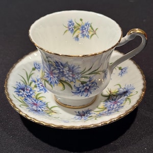 Puede incluir: Un juego de taza y platillo blancos con un diseño floral. La taza y el platillo están decorados con flores azules y hojas verdes. Los bordes están adornados con oro. La taza tiene un asa curva.