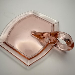 May include: A pink glass ashtray with a handle. The ashtray has a square shape with rounded corners and a clear rim. The handle is a loop shape. The glass has a slight pink hue.