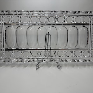 May include: A rectangular, clear glass serving tray with a decorative pattern around the edges and seven oval-shaped compartments. The tray is supported by a silver-colored metal stand, suitable for serving appetizers or desserts.