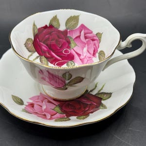 Puede incluir: Juego de taza y platillo blancos con un diseño de rosas en tonos rojos y rosas. La taza tiene un asa curvada y ribete dorado. El platillo tiene un borde festoneado y también presenta el diseño de rosas. Un juego de té clásico.
