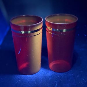 May include: Two red and gold drinking glasses. Each glass has a gold band around the middle. The glasses are cylindrical and appear to be made of glass or a similar transparent material. The background is dark blue.