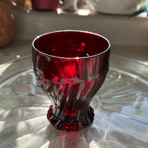 May include: A deep red glass with a textured, swirling pattern. The glass has a flared base and a wide opening. The glass is on a clear glass plate. The glass is likely for drinking.