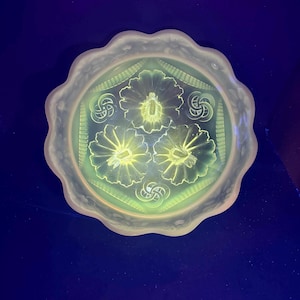 Puede incluir: Un plato redondo de vidrio opalino con borde festoneado. El plato presenta un diseño central de tres flores estilizadas y patrones geométricos decorativos. El vidrio tiene un aspecto suave y brillante, con un toque de verde.