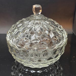 Puede incluir: Un bombonera de cristal transparente con un patrón de diamantes y forma redonda. El plato tiene una tapa con un pequeño pomo redondeado. El cristal refleja la luz, creando un efecto brillante. El plato es probablemente vintage.