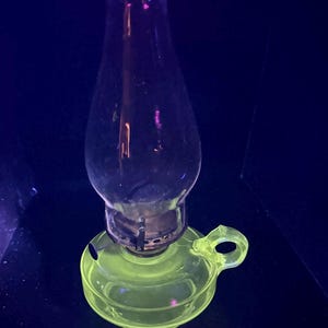 Könnte beinhalten: Eine Vintage-Öllampe aus klarem Glas mit einem gelb-grünen Sockel und Griff. Der Schornstein der Lampe ist klar, und der Sockel hat ein kreisförmiges Design. Die Lampe ist wahrscheinlich aus Pressglas und ein dekorativer Gegenstand.
