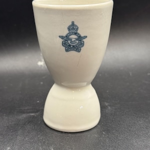 May include: Cream-coloured ceramic egg cup with a flared top and a rounded base. The cup features a blue emblem of the Royal Air Force. A vintage item.