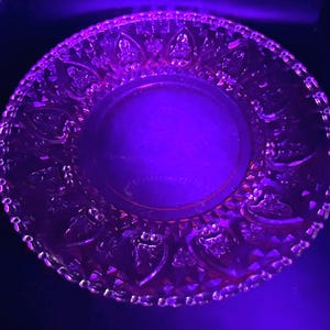 May include: A clear, decorative glass plate with an embossed pattern. The plate has a scalloped edge and a smooth central area. The glass reflects a purple hue, highlighting the intricate design.