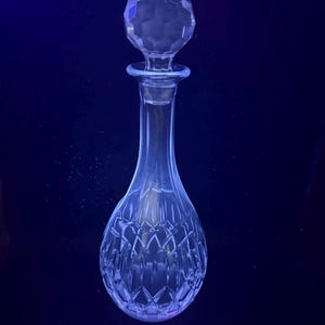 Puede incluir: Una jarra de cristal transparente con tapón facetado. La jarra tiene un cuello largo y una base bulbosa con un patrón tallado en diamante. El tapón tiene forma redonda y facetada. La jarra está diseñada para contener líquidos.