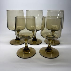 Può includere: Cinque bicchieri da vino vintage color ambra. Ogni bicchiere presenta una coppa arrotondata e uno stelo con base circolare. I bicchieri sono disposti su una superficie bianca, mostrando il loro design e colore unici.