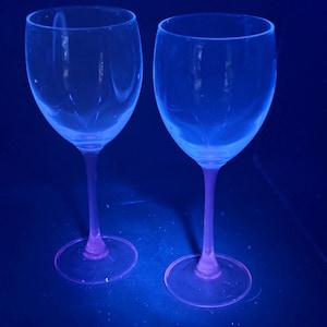 Könnte beinhalten: Zwei klare Weingläser mit rosa Stielen und Böden vor dunklem blauem Hintergrund. Die Gläser sind leer und aus Glas. Die Beleuchtung erzeugt einen Leuchteffekt. Geeignet zum Servieren von Wein.
