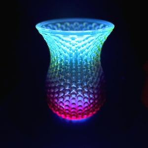 May include: A glowing glass vase with a textured, bubble-like pattern. The vase transitions in color from blue at the top, through green and yellow, to a vibrant pink at the base. The background is dark.