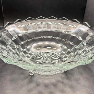 Puede incluir: Un cuenco de vidrio transparente con un patrón de diamantes y un borde festoneado. El cuenco tiene tres patas pequeñas y un centro decorativo. El cuenco se utiliza probablemente para servir fruta o aperitivos.