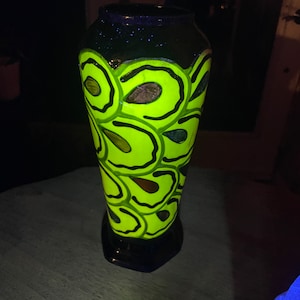 以下が含まれることがあります： 緑と黒の花柄の黒いセラミックの花瓶。花瓶は、様式化された花と葉のパターンで飾られています。花瓶はブラックライトで照らされ、緑色の塗料が光ります。