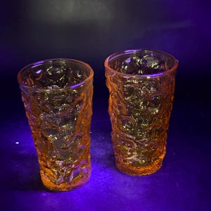May include: Two amber-coloured drinking glasses with a textured, organic pattern. The glasses are cylindrical and appear to be made of glass. The background is a dark purple.