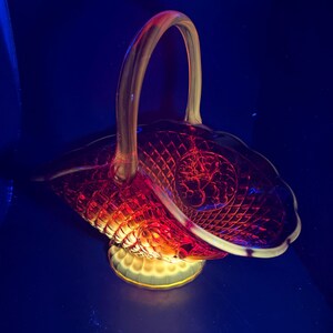 Puede incluir: Una cesta decorativa de cristal con asa. La cesta es de color rojo oscuro con un patrón de diamantes y un borde festoneado. La base es de color amarillo claro con un ribete dorado.