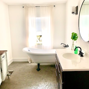 Puede incluir: Un ba&ntilde;o moderno y luminoso con una ba&ntilde;era blanca con patas de garra, un tocador oscuro con encimera blanca y un gran espejo redondo. La habitaci&oacute;n tiene una ventana con cortinas transparentes y suelo de hormig&oacute;n.