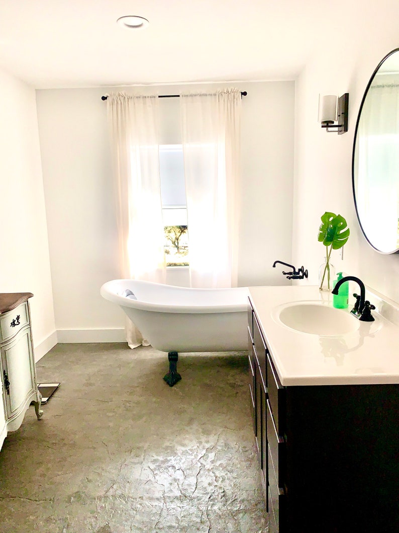 Puede incluir: Un ba&ntilde;o moderno y luminoso con una ba&ntilde;era blanca con patas de garra, un tocador oscuro con encimera blanca y un gran espejo redondo. La habitaci&oacute;n tiene una ventana con cortinas transparentes y suelo de hormig&oacute;n.