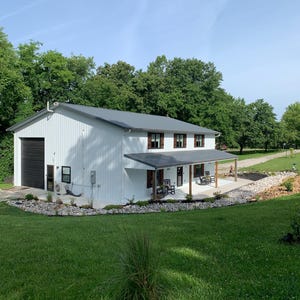 Puede incluir: Un edificio blanco de dos pisos con un techo de metal gris y una puerta de garaje negra. El edificio tiene un porche con mecedoras y est&aacute; rodeado de hierba verde y &aacute;rboles. El cielo es azul.