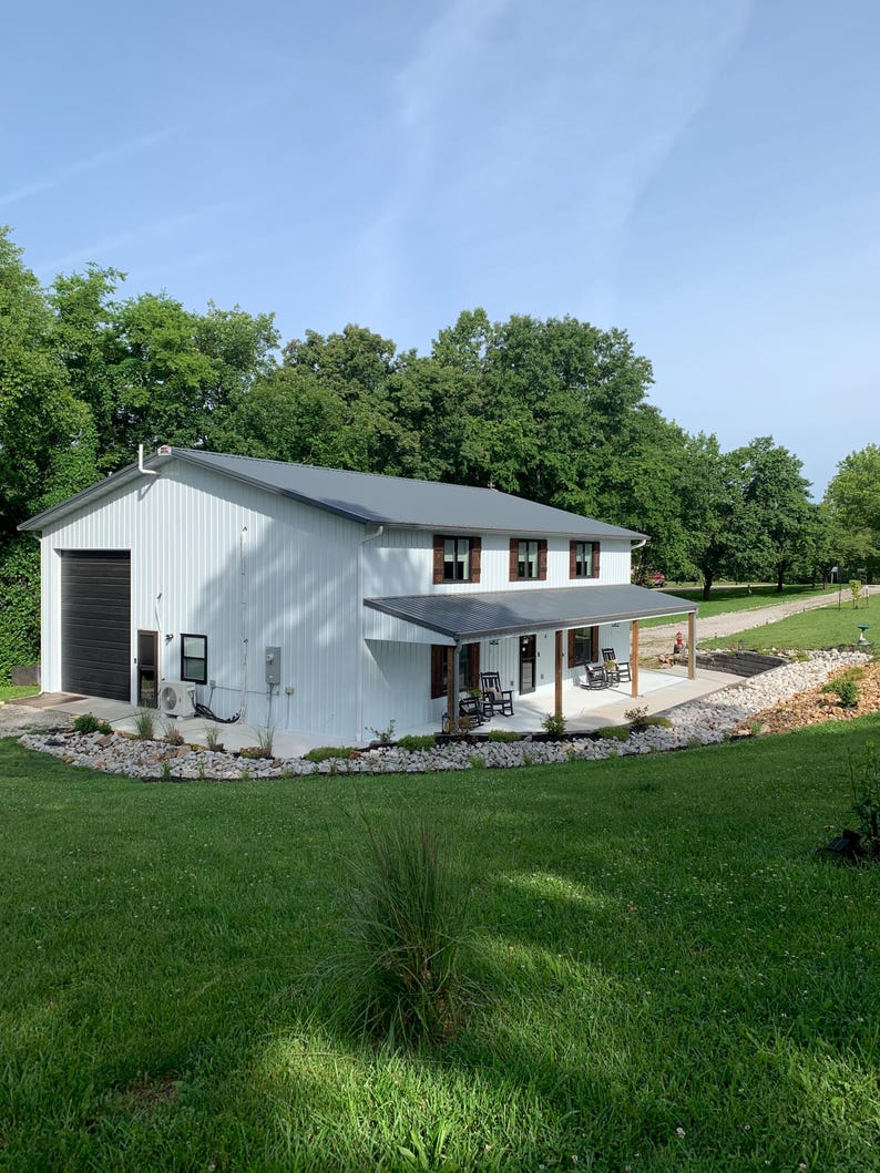 Puede incluir: Un edificio blanco de dos pisos con un techo de metal gris y una puerta de garaje negra. El edificio tiene un porche con mecedoras y est&aacute; rodeado de hierba verde y &aacute;rboles. El cielo es azul.