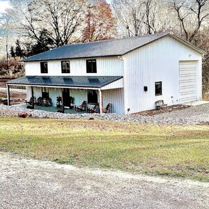 Puede incluir: Un edificio blanco de dos pisos con un tejado gris oscuro y un porche. El edificio tiene una puerta de garaje en el lado derecho. El porche tiene varias sillas y un tejado oscuro. El edificio est&aacute; rodeado de &aacute;rboles y hierba.