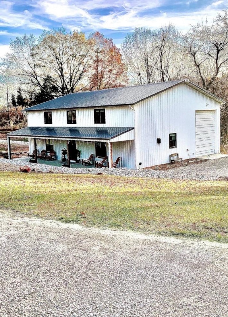 Puede incluir: Un edificio blanco de dos pisos con un tejado gris oscuro y un porche. El edificio tiene una puerta de garaje en el lado derecho. El porche tiene varias sillas y un tejado oscuro. El edificio est&aacute; rodeado de &aacute;rboles y hierba.