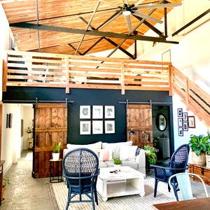 Puede incluir: Un amplio sal&oacute;n con techo alto, vigas de madera y un gran ventilador de techo. La habitaci&oacute;n cuenta con un &aacute;rea de loft con barandilla de madera, un sof&aacute; gris y dos sillas de mimbre azul marino. Puertas de granero de madera y obras de arte enmarcadas adornan la pared negra.