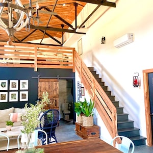 Puede incluir: Vista interior de un espacio habitable moderno y di&aacute;fano, con un techo alto revestido de madera y vigas a la vista. Una escalera de madera conduce a una zona de loft. Una gran mesa de madera est&aacute; en primer plano. Un candelabro cuelga del techo.