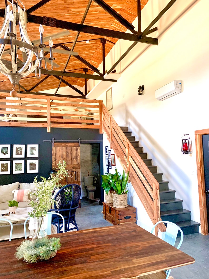 Puede incluir: Vista interior de un espacio habitable moderno y di&aacute;fano, con un techo alto revestido de madera y vigas a la vista. Una escalera de madera conduce a una zona de loft. Una gran mesa de madera est&aacute; en primer plano. Un candelabro cuelga del techo.