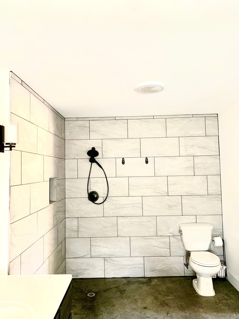 Puede incluir: Un ba&ntilde;o moderno con una ducha a ras de suelo con paredes de azulejos grises claros y un cabezal de ducha negro. Un inodoro blanco y un suelo de hormig&oacute;n gris oscuro completan el espacio. La habitaci&oacute;n est&aacute; bien iluminada.