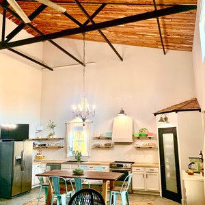 Puede incluir: Una cocina espaciosa con techo alto y vigas de madera a la vista. La habitaci&oacute;n cuenta con una gran mesa de comedor de madera con sillas turquesas, un refrigerador de acero inoxidable y gabinetes blancos. Una l&aacute;mpara de ara&ntilde;a cuelga sobre la mesa.