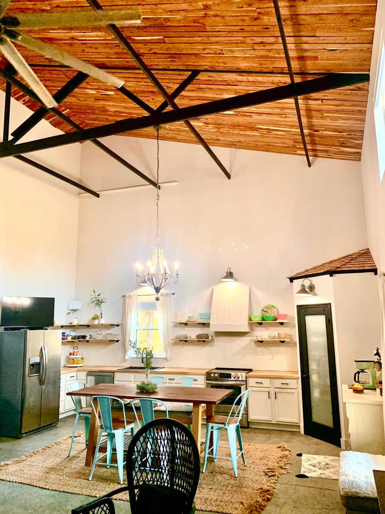 Puede incluir: Una cocina espaciosa con techo alto y vigas de madera a la vista. La habitaci&oacute;n cuenta con una gran mesa de comedor de madera con sillas turquesas, un refrigerador de acero inoxidable y gabinetes blancos. Una l&aacute;mpara de ara&ntilde;a cuelga sobre la mesa.