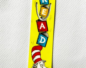 Puede incluir: Marcapáginas amarillo con el Gato en el sombrero. La palabra "READ" se deletrea con ilustraciones de libros coloridos. El marcapáginas tiene un agujero en la parte superior para una cuerda.